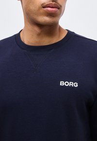 Κοντινό πλάνο ενός ατόμου που φοράει μπλε ναυτικό φούτερ με στρογγυλή λαιμόκοψη και το λογότυπο "BORG" τυπωμένο σε λευκό στο στήθος.