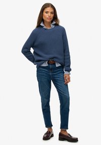 Blauwe gebreide trui met een relaxte fit over een wit gestreept shirt, gecombineerd met blauwe jeans en bordeauxkleurige loafers. Zachte texturen en casual stijl.