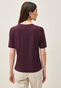 Burgundy triko s kulatým výstřihem, krátkými rukávy a hladkým povrchem. Nošeno s světlými kalhotami, které ukazují jednoduchý design.