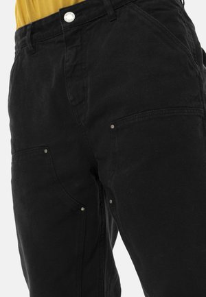 Pantalon cargo noir avec poches renforcées et détails à rivets, porté avec un haut jaune moutarde rentré, visible de la taille à mi-cuisse.