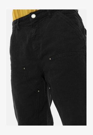 Pantalon cargo noir avec poches renforcées et détails à rivets, porté avec un haut jaune moutarde rentré, visible de la taille à mi-cuisse.