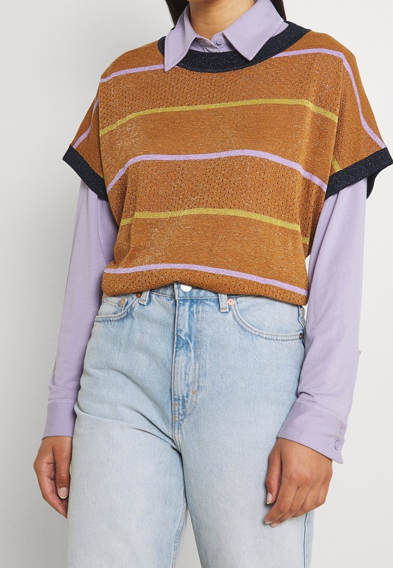 T-shirt à manches courtes, en tricot marron avec des rayures violettes et jaunes, porté sur une chemise à manches longues lavande ; associé à un jean bleu clair.