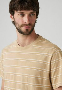 Beżowa bawełniana koszulka z poziomymi białymi i jasnopomarańczowymi paskami, z okrągłym dekoltem i krótkimi rękawami. Miękka tekstura.