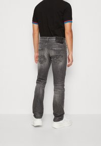 Jeans de mezclilla gris con un diseño desgastado, estilo de cinco bolsillos, bolsillos traseros cosidos y una etiqueta de cuero negro en la cintura. Zapatillas blancas puestas.