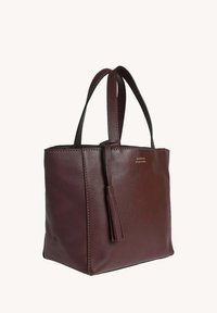 Sac fourre-tout en cuir bordeaux avec poignées doubles, finition texturée et accent en pompon. Forme carrée, design robuste, matériel minimal.