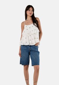 Weißes, schichtweises ärmelloses Oberteil mit goldenen Verzierungen, kombiniert mit Jeansshorts. Das Oberteil hat einen strukturierten Stoff und eine lockere Silhouette.
