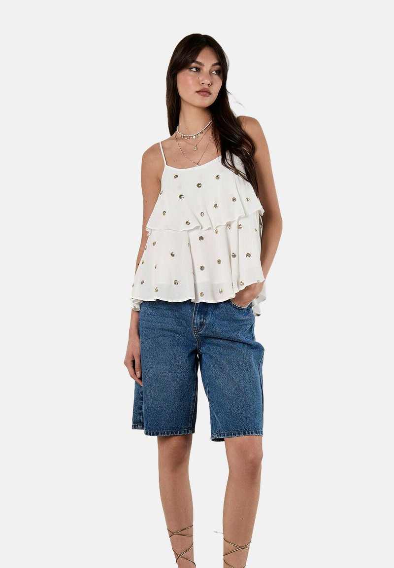 Weißes, schichtweises ärmelloses Oberteil mit goldenen Verzierungen, kombiniert mit Jeansshorts. Das Oberteil hat einen strukturierten Stoff und eine lockere Silhouette.