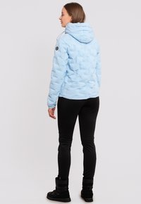 Helle blaue Pufferjacke mit Kapuze, mit einer Stepptextur, verstellbarem Kordelzug und einem schwarzen Emblem am Ärmel. Schwarze Leggings und Stiefel runden den Look ab.