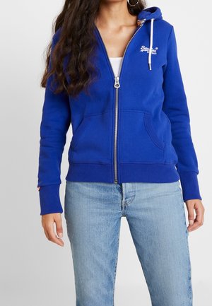 Kobieta ubrana w niebieską bluzę z zamkiem i białymi sznurkami oraz logo na piersi, w połączeniu z jasnoniebieskimi dżinsami na jednolitym tle.