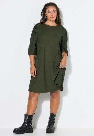 Junge Frau mit langen Haaren, die ein lockeres olivgrünes Kleid und schwarze, mit Nieten besetzte Kampfstiefel vor einem schlichten weißen Hintergrund trägt.