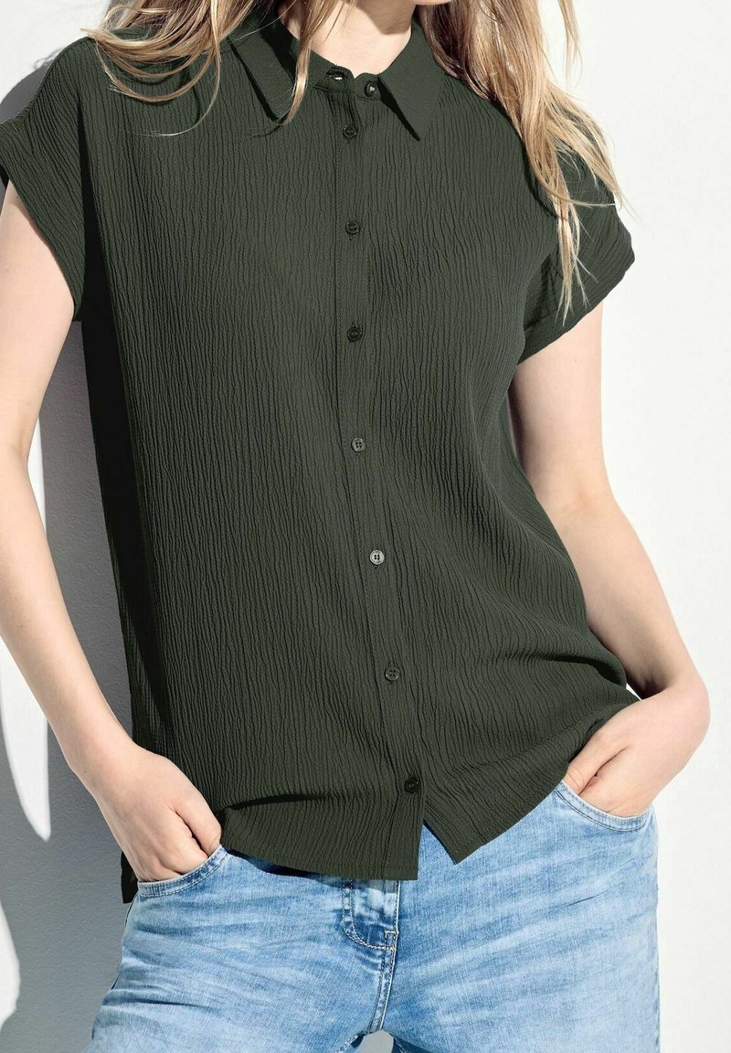 Femme portant une chemise à manches courtes texturée vert foncé boutonnée, avec un jean bleu clair, les mains reposant dans les poches avant.