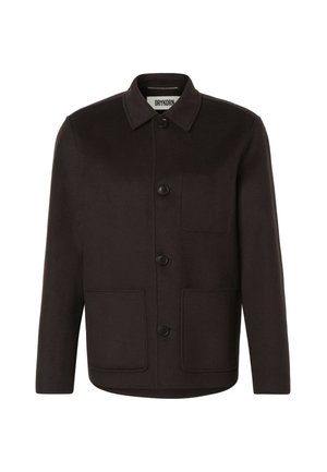 Homme debout les mains dans les poches, portant une veste marron foncé boutonnée, un pantalon beige à jambes larges et des chaussures noires sur un fond uni.