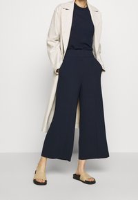 Marinblå vida byxor med struktur, hög midja och fickor, i kombination med en ljusbeige lång kappa och bruna sandaletter på en neutral bakgrund.