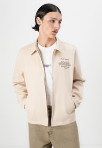 Jachetă bej ușoară cu fermoar frontal și manșete elastice. Prezintă text brodat în brun, purtată deasupra unei tricouri albe și a unor pantaloni bej deschis.