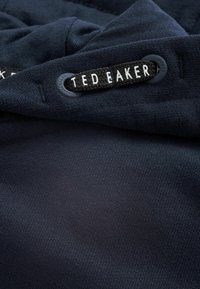 Niebieska tkanina w marynarskim odcieniu z teksturowanym wykończeniem, z czarną metką i białym napisem "TED BAKER" oraz pasującym plastikowym akcentem.
