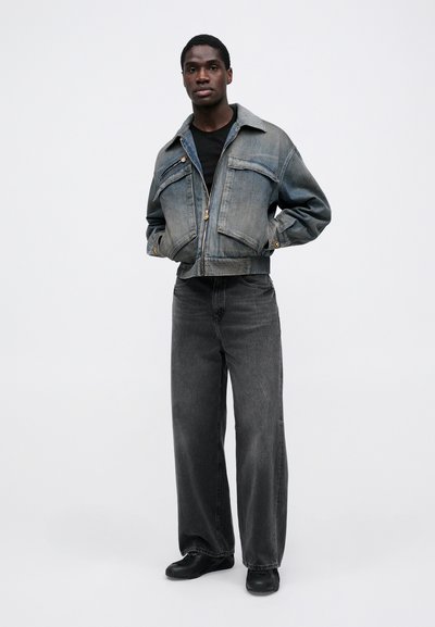 Cropped denim jacket in light blue with two chest pockets, paired with high-waisted, loose-fitting black jeans and black shoes.