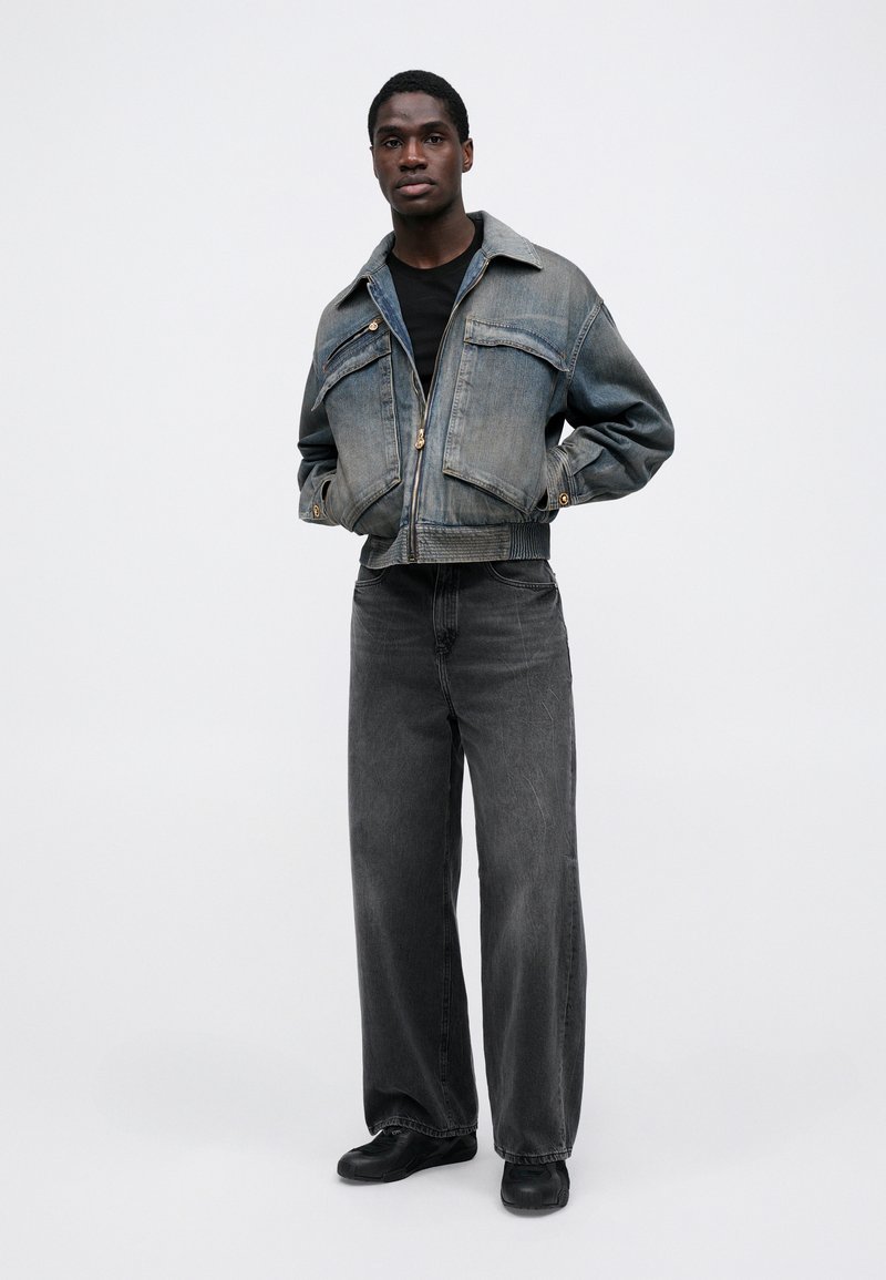 Gekürzte Jeansjacke in Hellblau mit zwei Brusttaschen, kombiniert mit hoch taillierten, locker sitzenden schwarzen Jeans und schwarzen Schuhen.