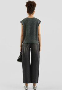 Woman with curly hair in a sleeveless grey sweater, wide-leg black jeans, patterned shoes, holding a small black handbag, facing away.