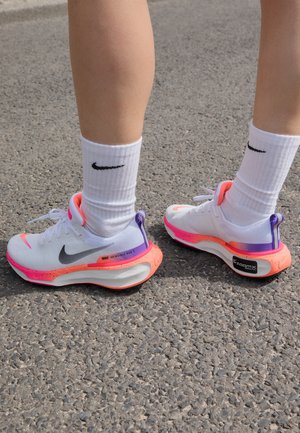 Person wearing white Nike Invincible Run 3 running shoes with pink and purple accents, standing on gray asphalt.