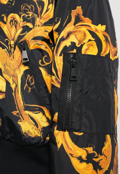 Veste bomber noire avec un motif floral orné jaune. Comprend des poches latérales zippées et des poignets côtelés. Texture lisse, semblable à du satin.