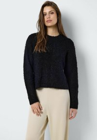 Zwarte fuzzy trui met lange mouwen en een ronde hals. Het heeft een korter ontwerp en een zachte textuur, gecombineerd met beige broek.