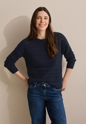Donkerblauwe trui met gestructureerde horizontale strepen en lange mouwen, gecombineerd met blauwe jeans. Model staat tegen een lichte muur.