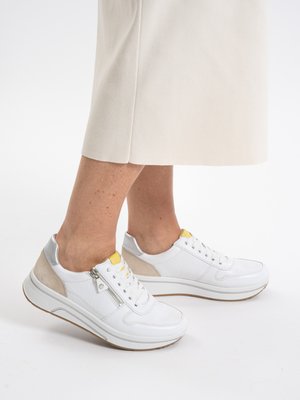 Baskets blanches avec des accents argentés et beiges, détail jaune sur la languette, fermeture éclair latérale, portées avec une jupe crème sur un fond blanc uni.