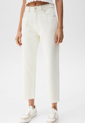 Femme portant un jean blanc cassé taille haute, jambes droites, ourlets bruts, et des baskets blanches, debout devant un fond uni.