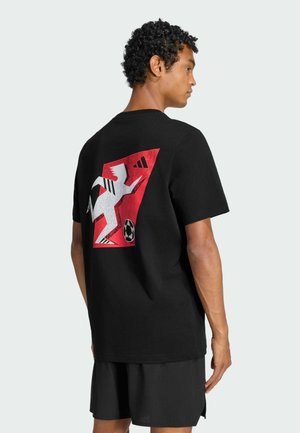 Homme vu de profil portant un t-shirt noir avec un graphique abstrait rouge et blanc représentant un joueur de football et un short noir sur fond uni.