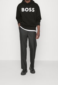 Sudadera de algodón negra con impresión blanca "BOSS", superpuesta sobre una camisa blanca, combinada con pantalones grises y zapatos negros de suela gruesa.