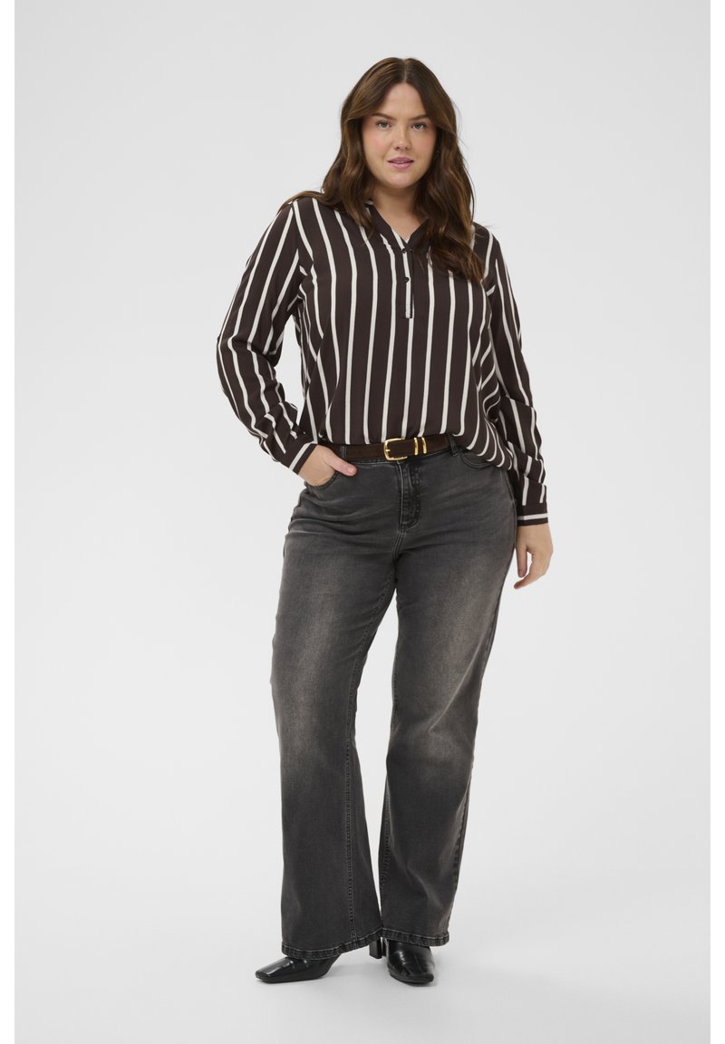 Femme debout, main dans la poche, portant un chemisier rayé noir et blanc, un jean noir délavé et des chaussures à talons noires sur un fond uni.