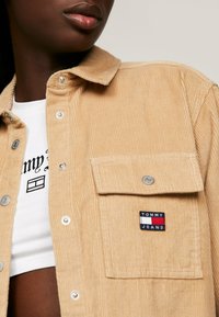 Camisa de pana en beige claro con botones a presión, un bolsillo frontal y un parche de marca "TOMMY JEANS". Debajo, se ve una camiseta gráfica blanca.