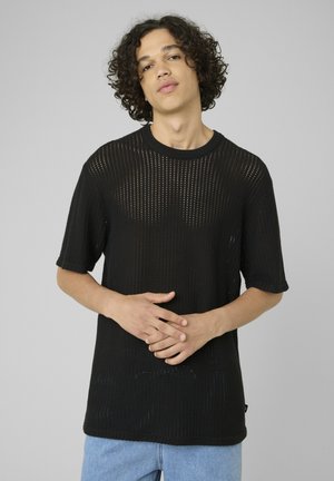 Jeune homme aux cheveux bouclés portant un t-shirt tricoté noir perforé à manches courtes et un jean bleu clair sur un fond uni.