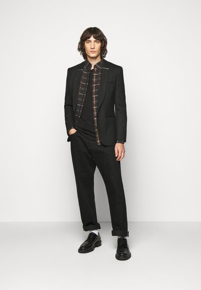 Black blazer with notch lapels, layered over a black and plaid checkered shirt, paired with black trousers and black shoes, against a white backdrop.