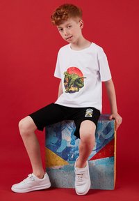 Garçon aux cheveux roux portant un t-shirt blanc avec un dinosaure et un short noir, assis sur un cube coloré sur un fond rouge.