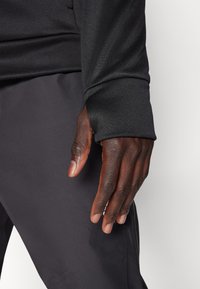 Parte superior deportiva de manga larga negra con puños ajustados, combinada con pantalones deportivos gris oscuro. Textura suave, diseño funcional para la ropa activa.