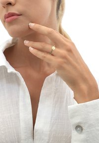 Goldring mit einem kreisförmigen Design, das einen zentralen Punkt zeigt, getragen an einer Hand mit neutralem Nagellack, vor einem weißen Hintergrund.