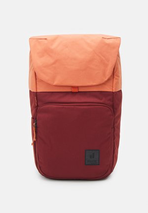 Sac à dos en tissu rouge et orange avec un rabat supérieur, une poche zippée à l'avant et un patch logo. Surface texturée avec des sections de couleur contrastante.