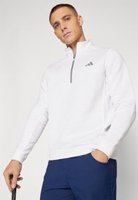 Maglietta sportiva bianca a maniche lunghe con colletto a mezza zip e texture a coste. Presenta un accento con logo grigio sul petto.