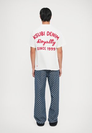 Hvit t-skjorte med stor rød tekst som leser "KSUBI DENIM Royalty SINCE 1999." Kombinert med blåmønstrede vide bukser.