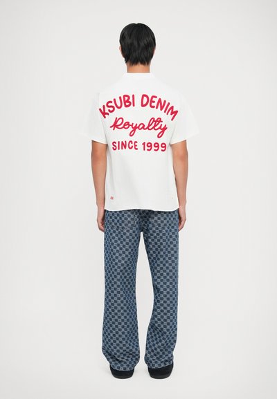 Fehér póló, melyen nagy piros betűkkel olvasható: "KSUBI DENIM Királyság 1999 óta." Kék mintás, bő szabású nadrággal párosítva.