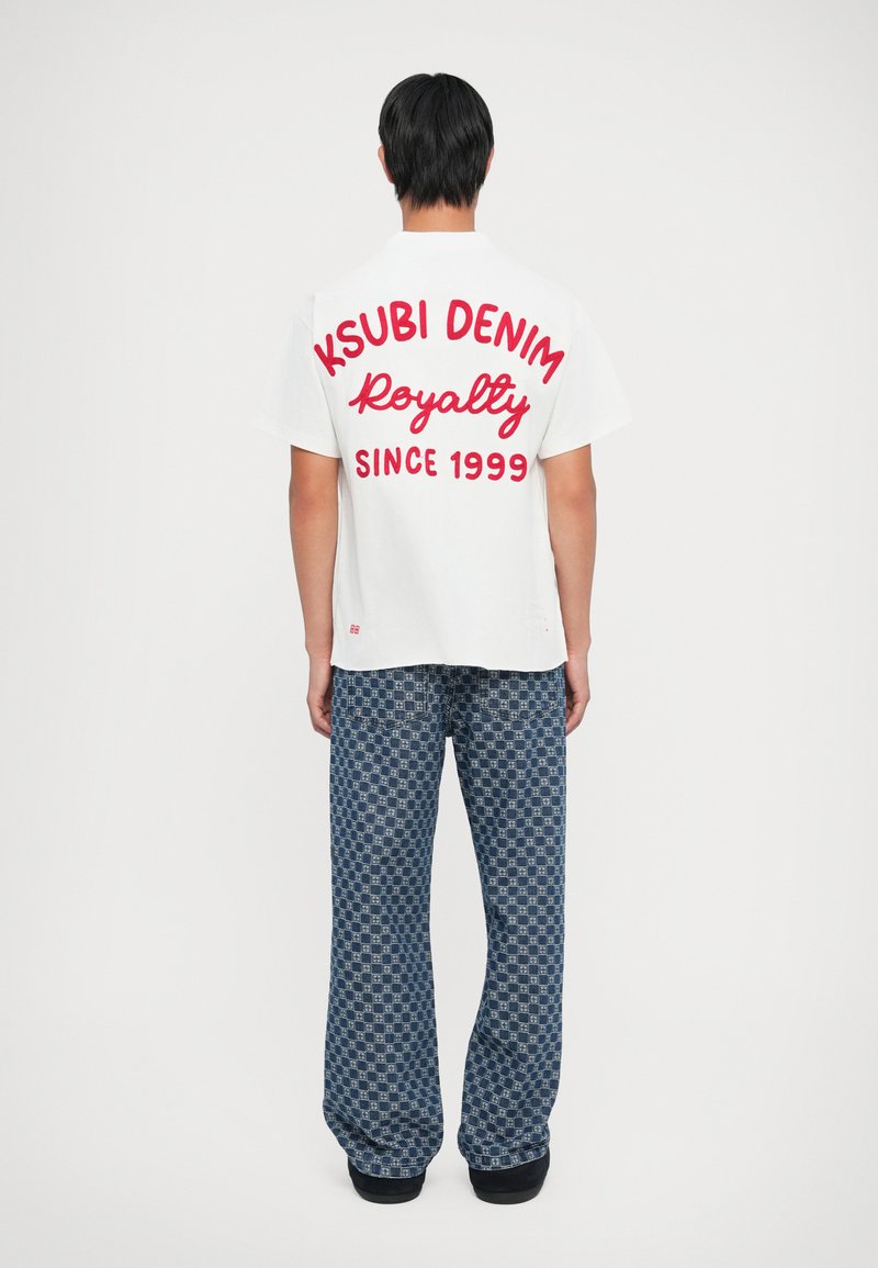 Balta t-kreklā ir liels sarkans uzraksts "KSUBI DENIM Royalty SINCE 1999." Apģērbs kombinēts ar zilām rakstainām platām biksēm.