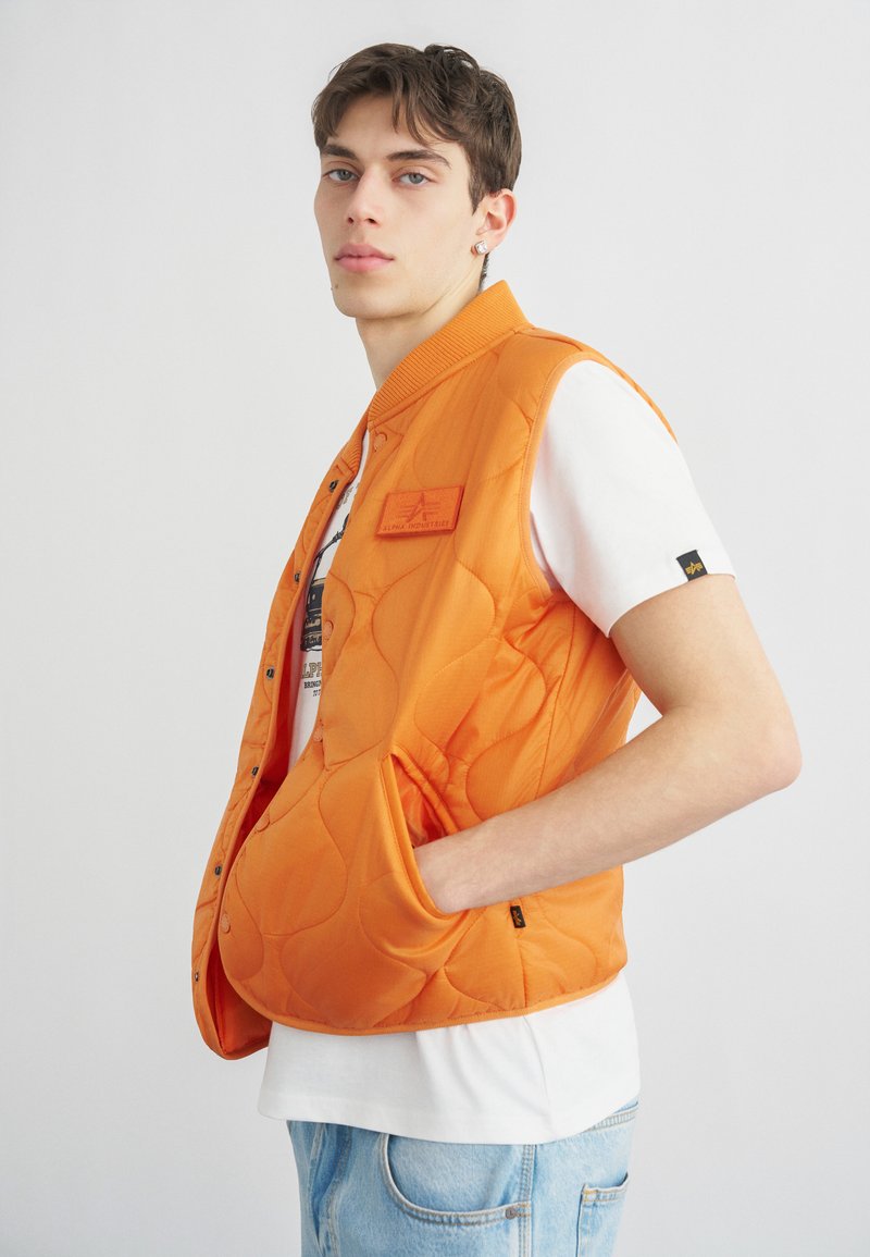 Jeune homme portant une veste matelassée orange vif sur une chemise blanche et un jean bleu clair, debout avec une main dans la poche de la veste.