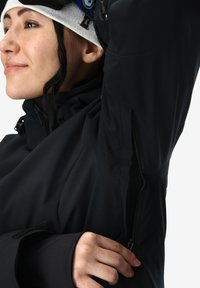 Giacca nera ad alte prestazioni con una presa d'aria con zip sotto le ascelle, polsini regolabili e cappuccio. Texture liscia con un design aderente.