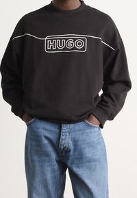 Sudadera negra con el logo "HUGO" bordado en blanco, cuello redondo acanalado y corte relajado, combinada con jeans de mezclilla azul claro.