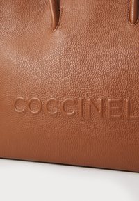 Sac à main en cuir marron avec une finition texturée et un logo en relief "COCCINELLE" sur le devant, au design minimaliste et comprenant deux poignées.