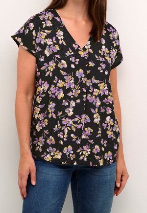 Kobieta w czarnej bluzce z dekoltem w serek i krótkimi rękawami, ozdobionej fioletowo-żółtym kwiatowym wzorem, zestawionej z niebieskimi dżinsami.