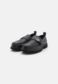 Sorte slip-on sko med en glat læderoverdel, syet detaljer og en bred strop over vristen med en metallisk accent. Struktureret sål.
