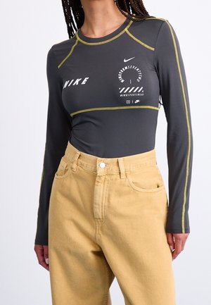 Kobieta w ciemnoszarej bluzce Nike z długim rękawem, żółtym przeszyciem i beżowych spodniach z wysokim stanem na jasnym tle.