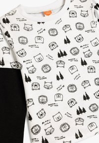 Weißes Baumwollhemd mit langen Ärmeln, das schwarze Drucke mit Tieren und Waldmotiven zeigt, darunter Bären, Löwen und Textakzente. Schwarze Ärmel-details.